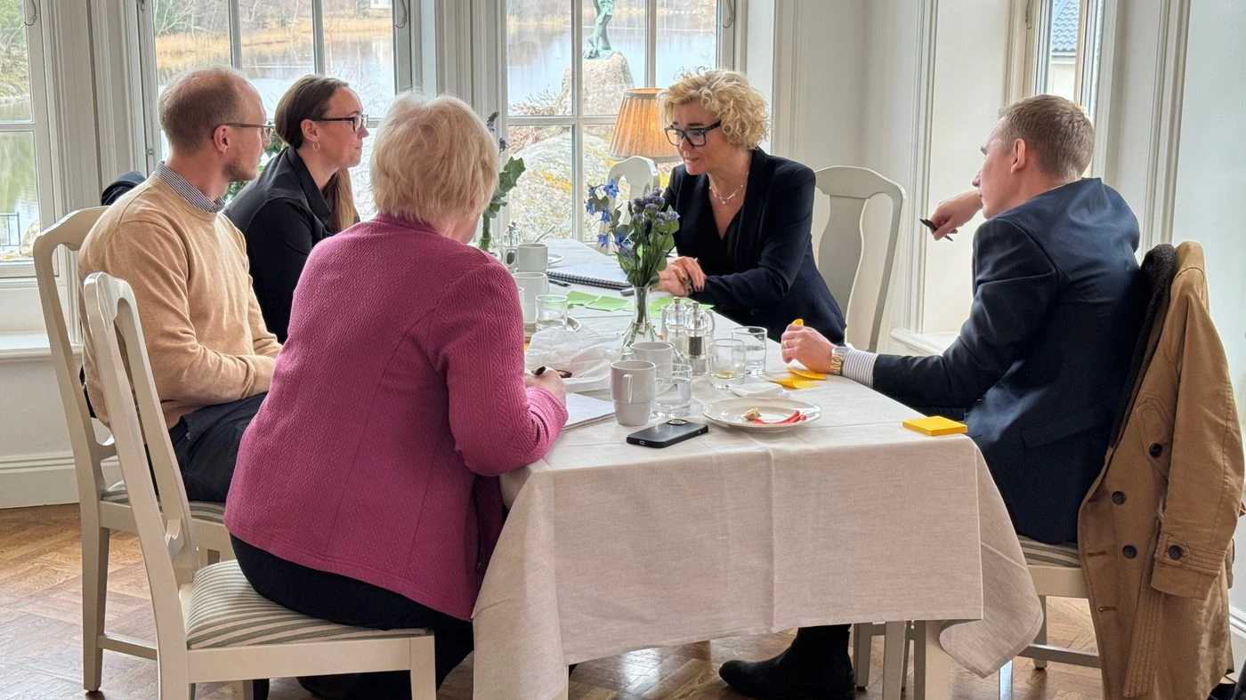 Fem personer i samtal runt ett dukat bord vid fönster mot en sjö